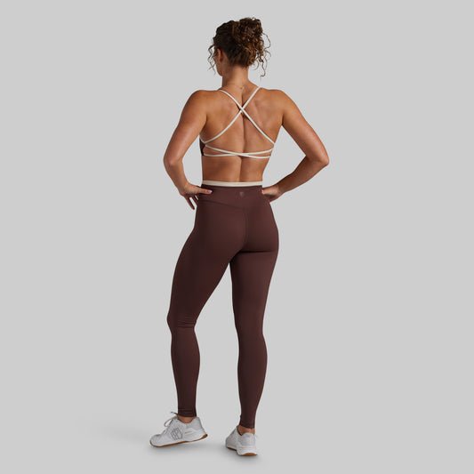 A woman shown from the back wearing a brown athletic leggings set with a matching sports bra featuring cream-colored crisscross straps, posed against a solid light gray background.