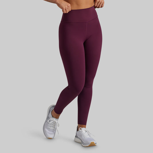 A model wearing high-waisted, full-length athletic leggings in a deep plum purple color, paired with white sneakers, shown from the waist down on a solid light gray background.