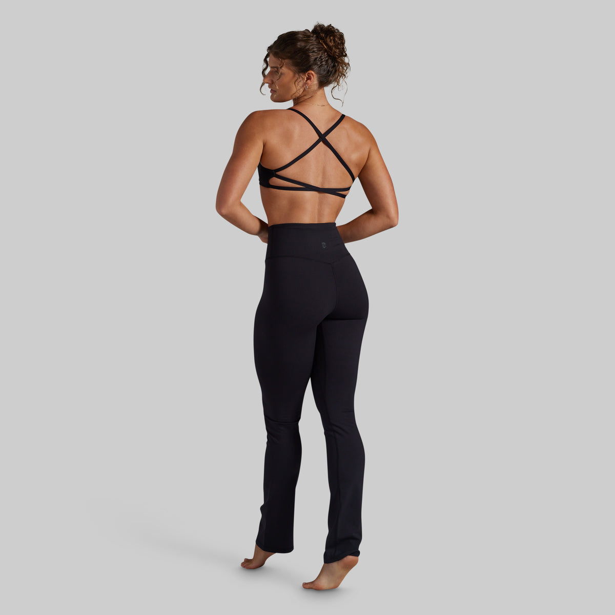 A woman is shown from the back wearing a black strappy athletic bra and black high-waisted yoga pants against a solid gray background.