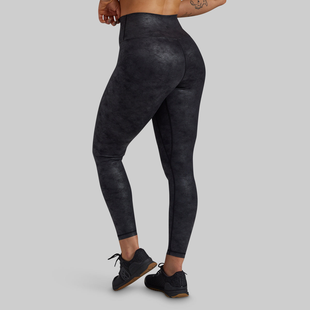 Rear three-quarter view of a model wearing high-waisted, full-length athletic leggings with a dark charcoal gray and black mottled texture, paired with black sneakers, against a solid light gray background.