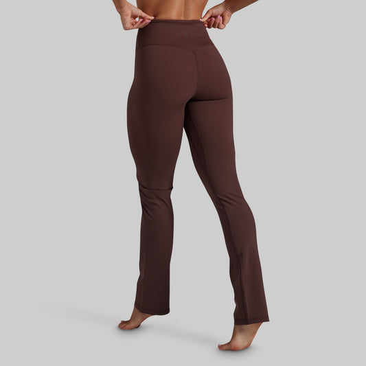 A person shown from the waist down wearing dark brown high-waisted yoga pants, standing barefoot against a solid light gray background.