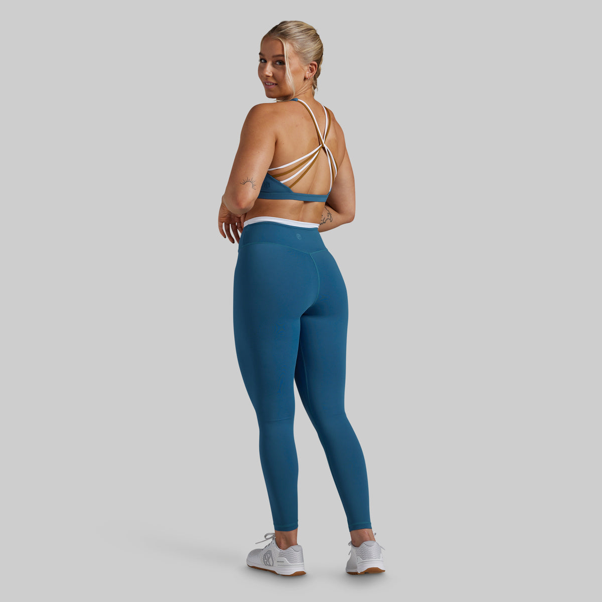 A woman modeling a matching athletic set consisting of a teal blue sports bra with beige straps and teal blue high-waisted leggings, shown from a rear three-quarter angle against a solid gray background.