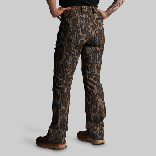 A person shown from the back wearing camouflage hunting pants, a black shirt, and brown boots against a solid gray studio background.