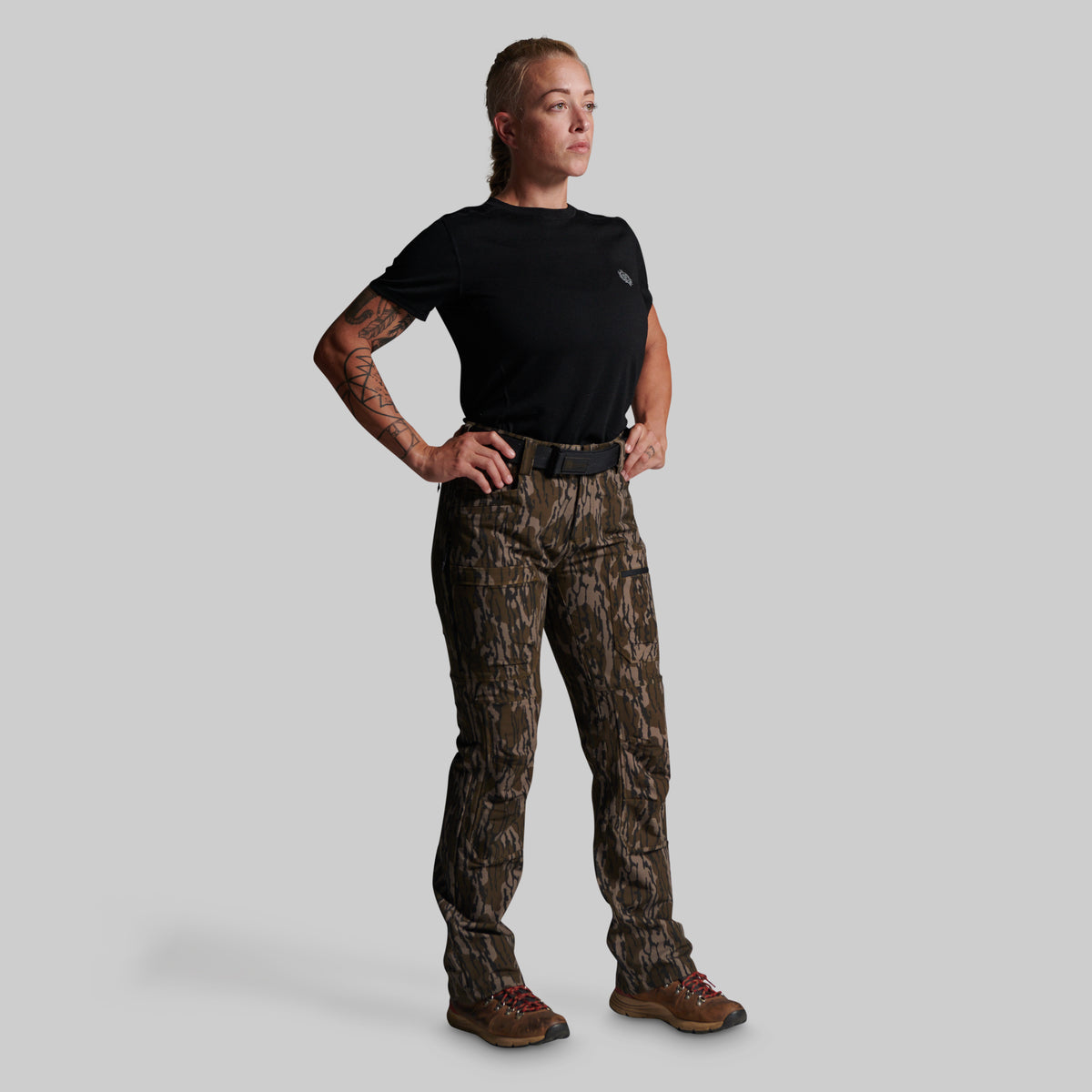 A woman modeling a pair of brown and black camouflage hunting pants, paired with a black t-shirt, black belt, and brown hiking boots, against a solid gray background.
