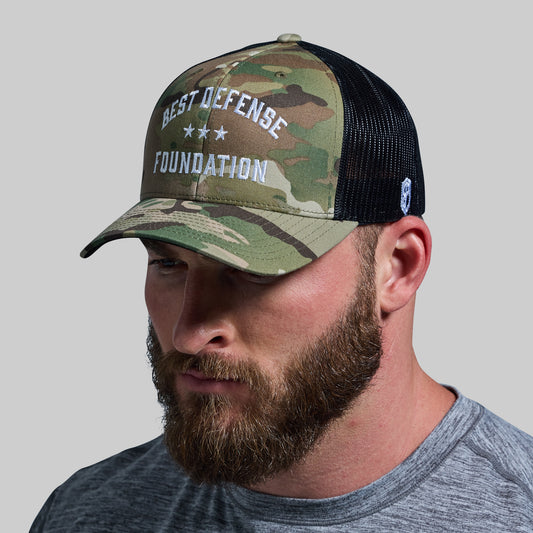 A person wearing a camouflage trucker hat with 'BEST DEFENSE FOUNDATION' text on the front.