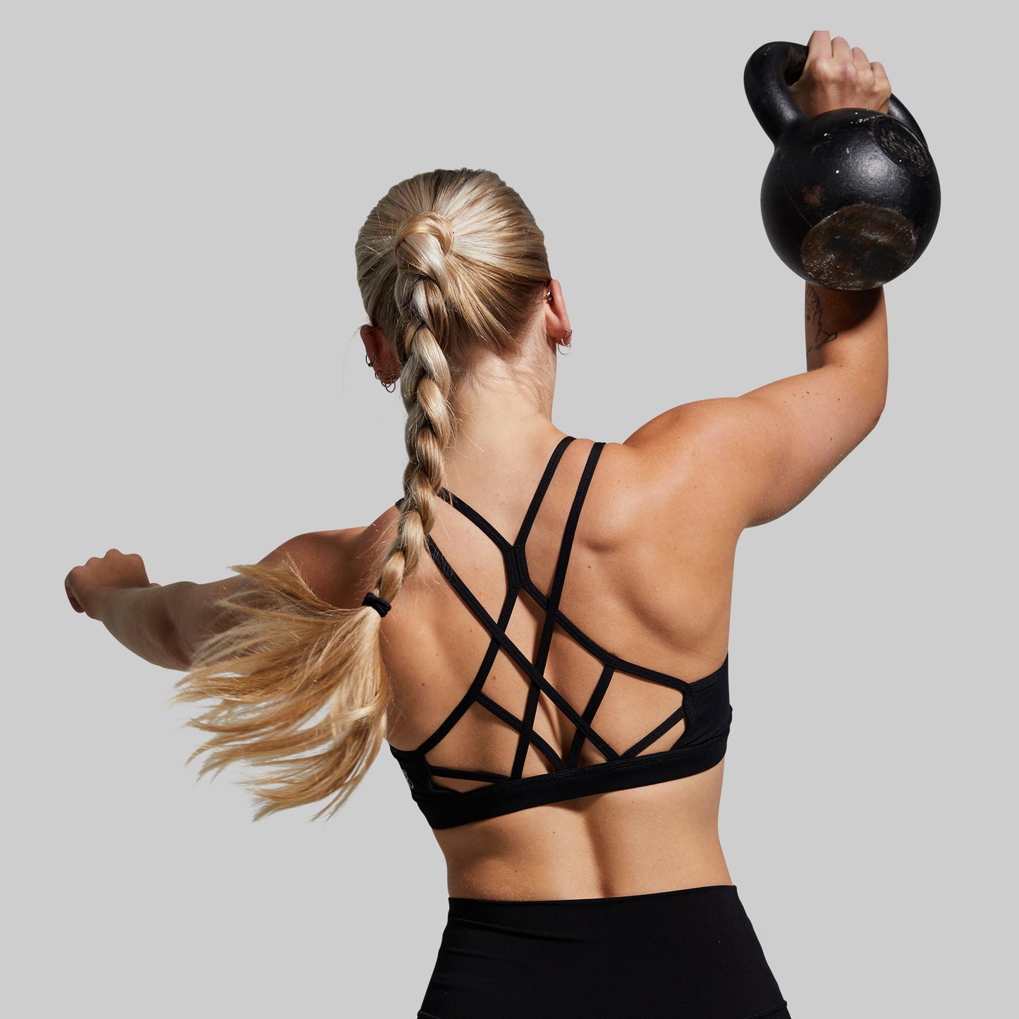 Woman wearing black strappy sports bra lifting a kettlebell with a plain background