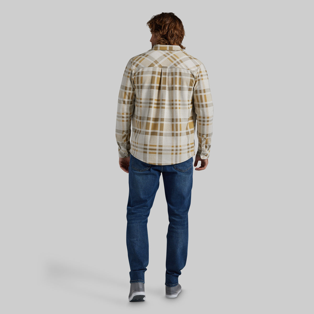 A man viewed from behind wearing a cream and tan plaid button-down shirt, blue jeans, and gray sneakers, standing against a solid light gray background.