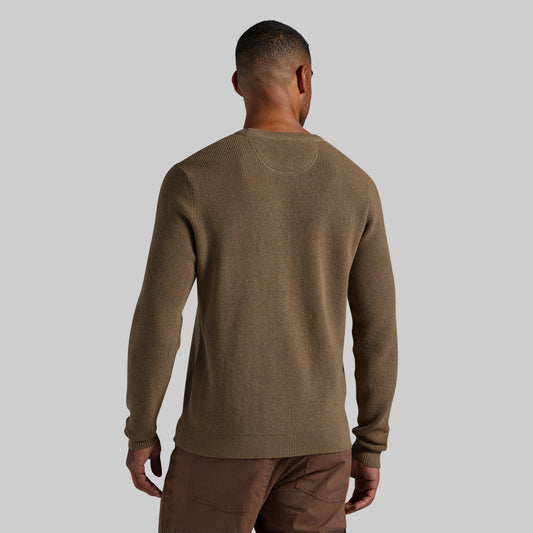 A man viewed from the back wearing a brown knit crewneck sweater and brown pants against a solid light gray background.