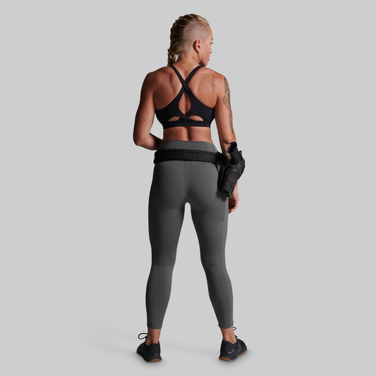 A woman shown from the back wearing dark gray full-length leggings and a black sports bra, standing against a light gray studio background. She is wearing a black belt with a holster