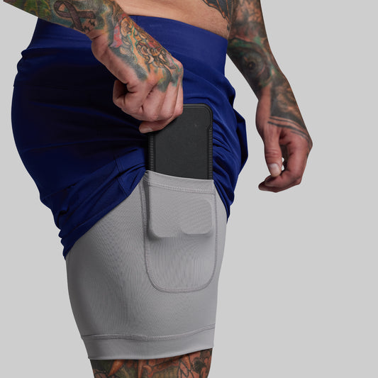 Person wearing a blue athletic shorts with gray liner with a pocket holding a phone, on a plain background