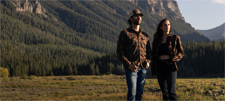 A man and a woman standing outdoors in a mountain meadow with forested mountains in the background. The man wears a brown hat, a brown plaid shirt, and blue jeans, while the woman wears a dark top and a dark plaid shirt with dark pants.