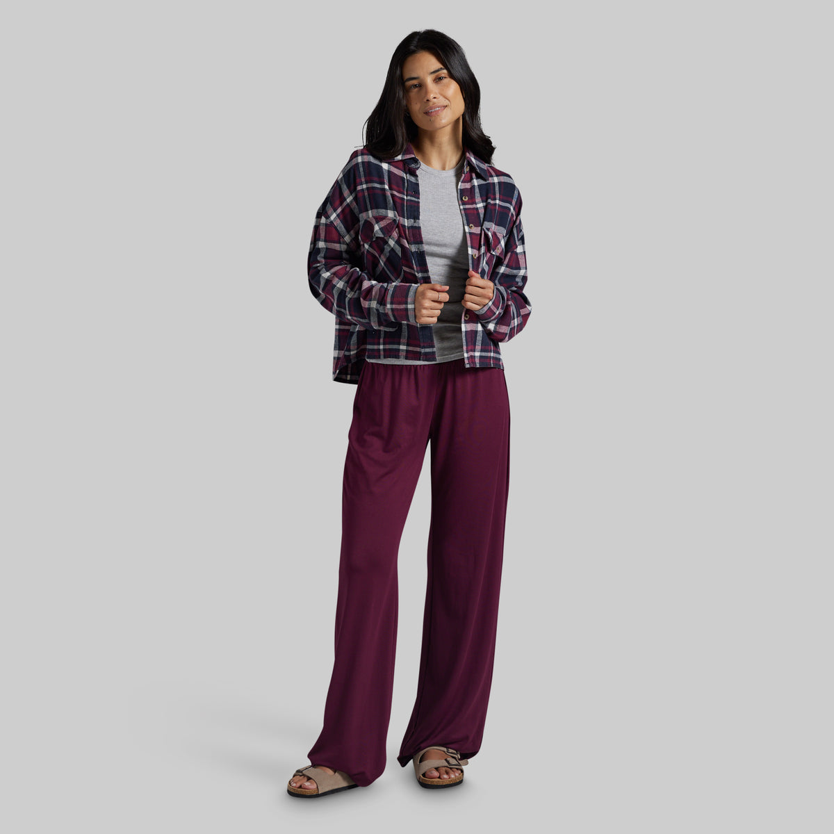 A woman modeling a two-piece lounge set consisting of a dark red and navy plaid flannel shirt and deep burgundy wide-leg pants, styled with a light gray t-shirt and brown sandals against a solid gray background.