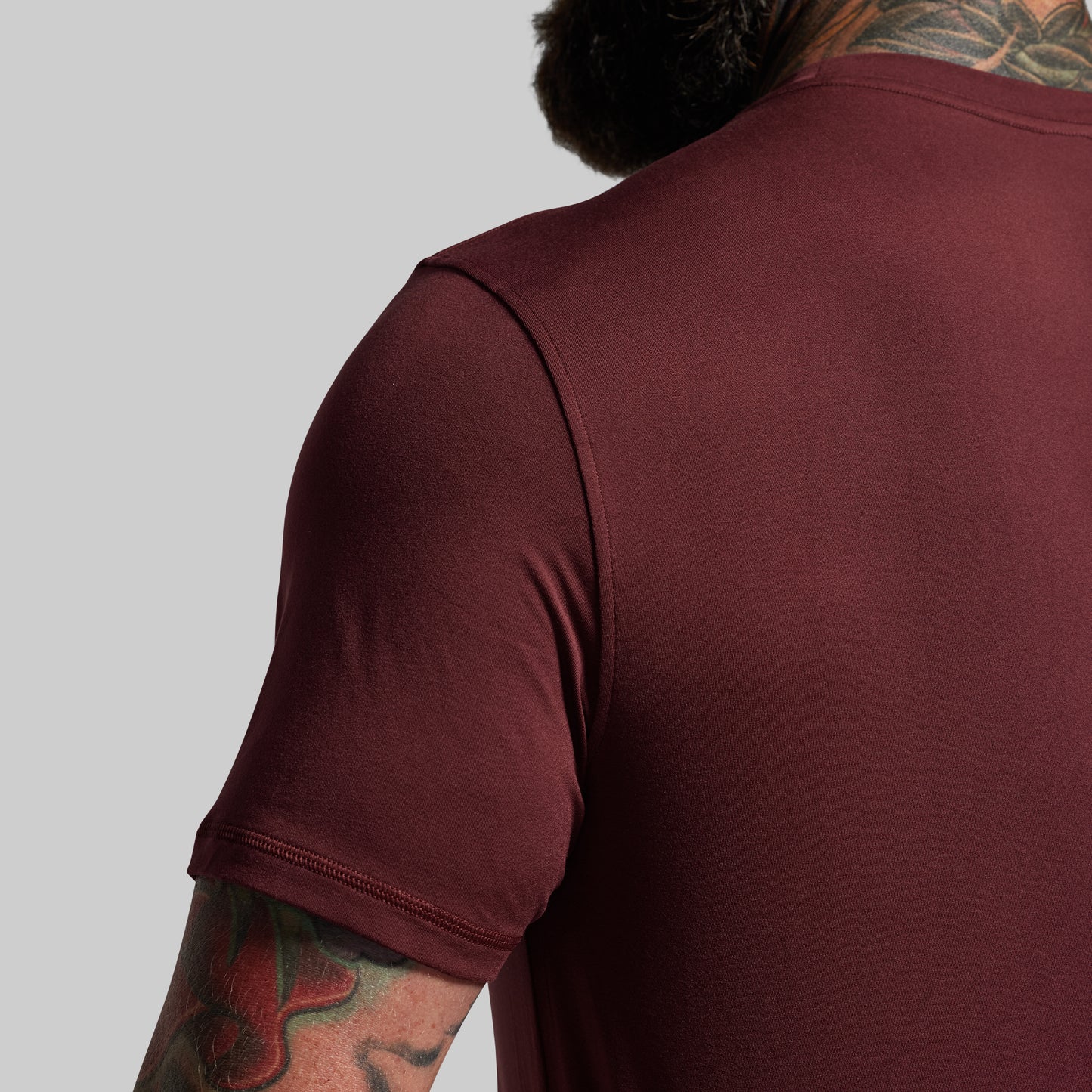 Person wearing a maroon t-shirt with tattoos on arms and neck against a gray background