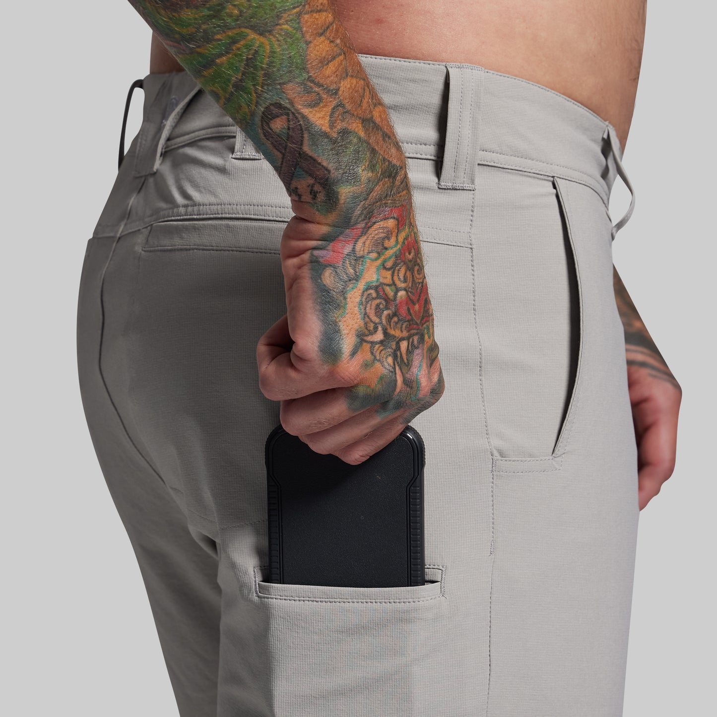 Person wearing light gray pants with a tattooed arm holding a black device.