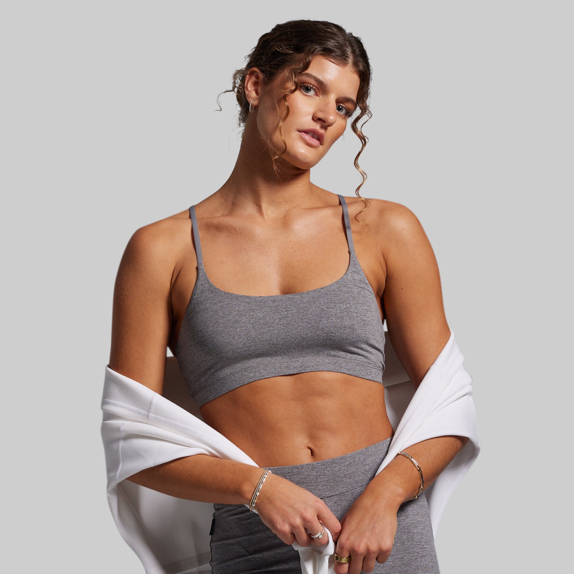Woman wearing a gray sports bra and leggings with a white towel draped over her shoulders on a light gray background.