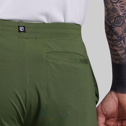 Close-up of green shorts with a brand logo on the waistband, white shirt, and tattooed arm.