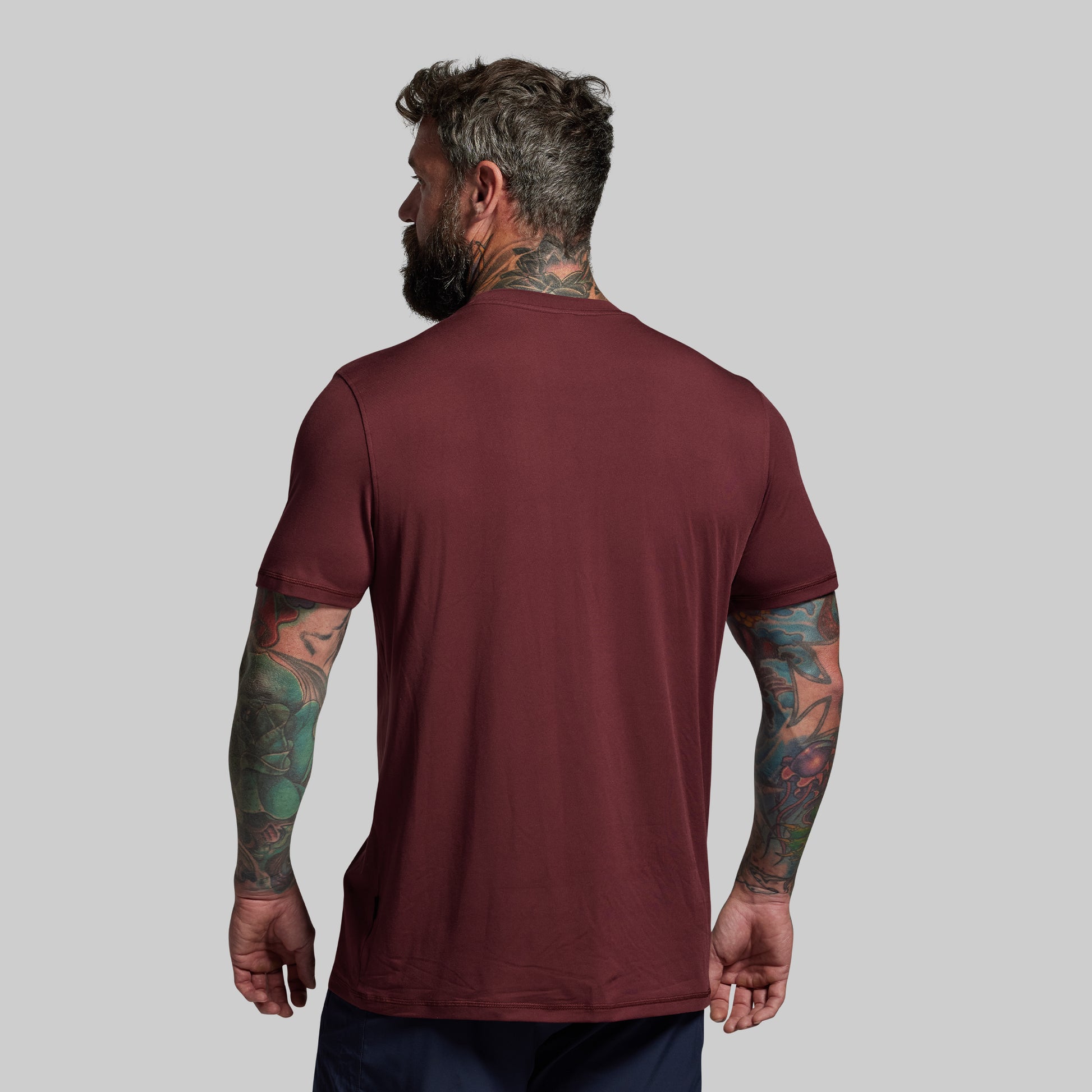 Man wearing a maroon t-shirt with visible tattoos on arms against a gray background