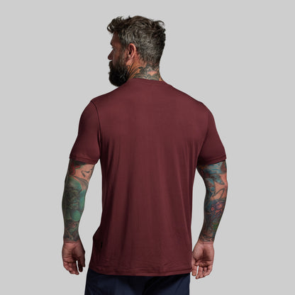 Man wearing a maroon t-shirt with visible tattoos on arms against a gray background