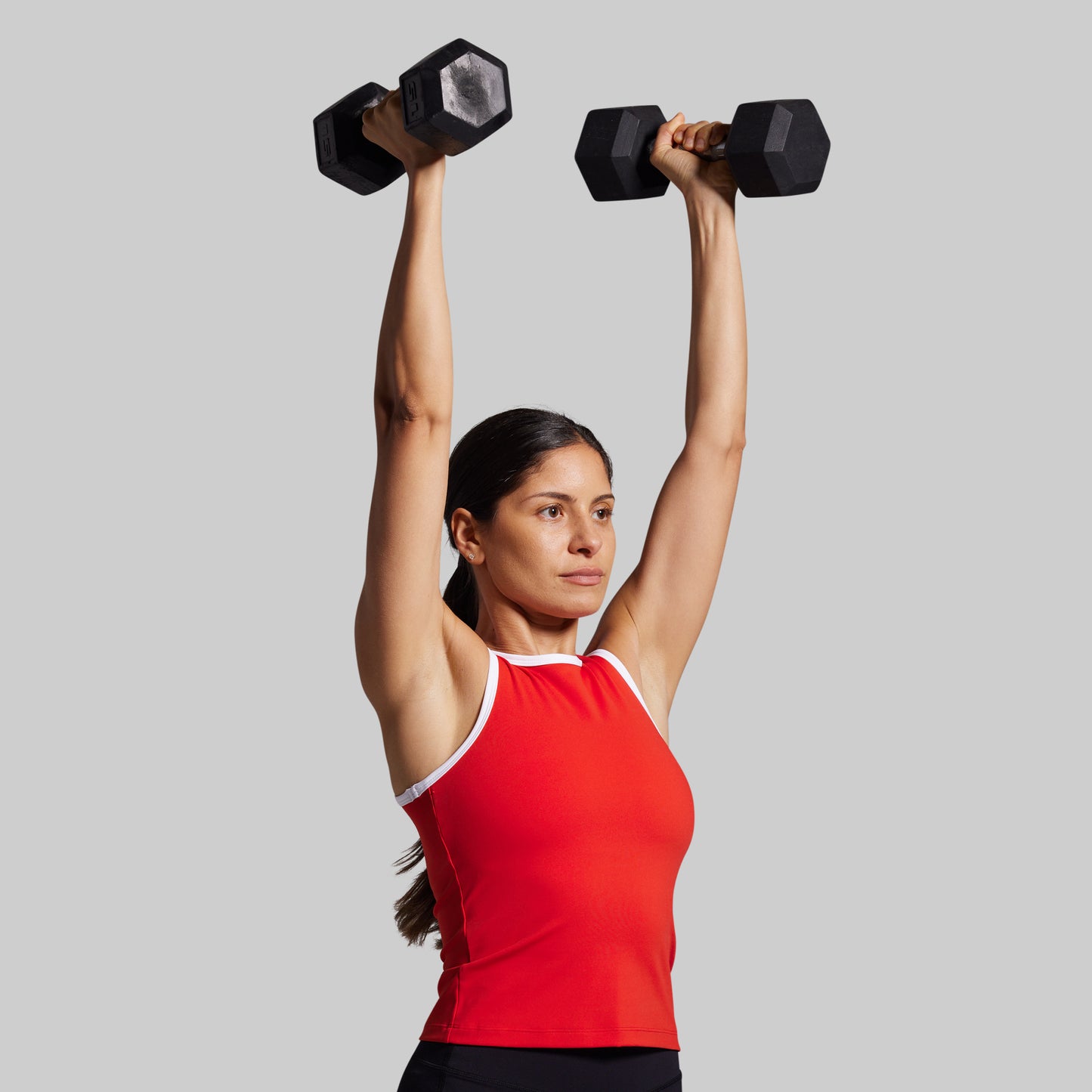 Woman lifting dumbbells above her head wearing a red tank top against a plain background