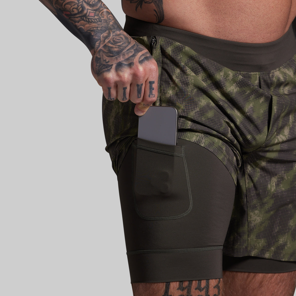 Person wearing a green and black camouflage shorts with a phone in a pocket on a gray background