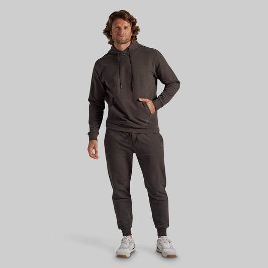 A man modeling a dark grey matching tracksuit, consisting of a pullover hoodie and jogger pants, paired with white sneakers, against a solid light gray background.