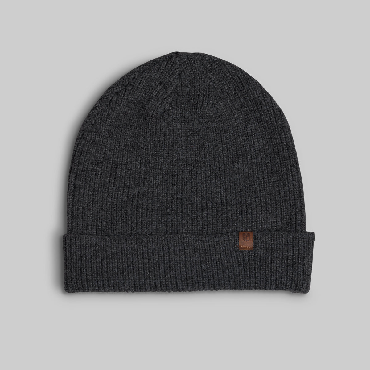 A dark charcoal gray ribbed knit beanie hat with a folded cuff and a small brown leather logo patch on the side, shown against a solid light gray background.