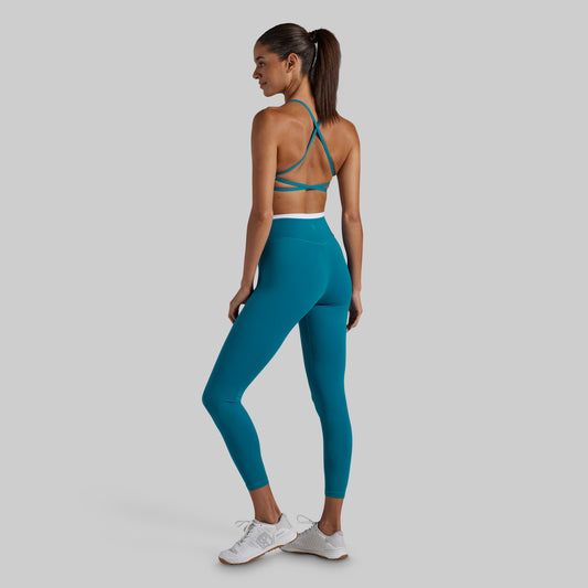 A woman wearing a teal sports bra and matching teal leggings with a white waistband, shown from the back against a solid gray background.