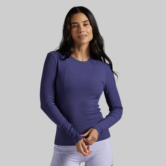 A woman modeling a long-sleeve crewneck athletic top in a solid periwinkle purple color, paired with white leggings, against a solid light gray background.