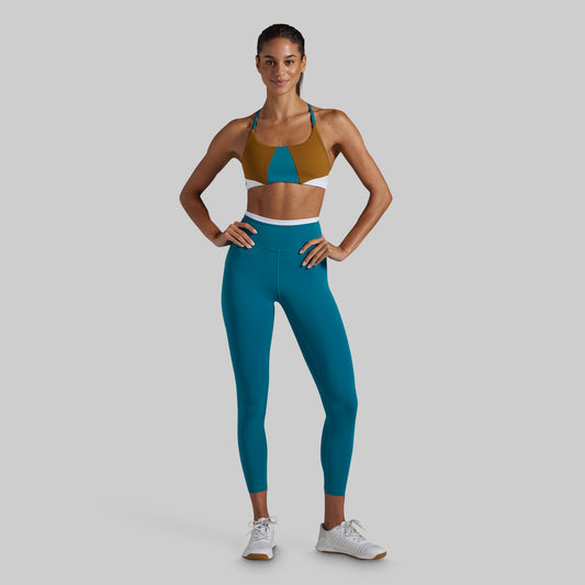 A woman modeling a two-piece activewear set consisting of a color-blocked sports bra in brown, teal, and white, paired with high-waisted teal leggings, against a solid gray background.