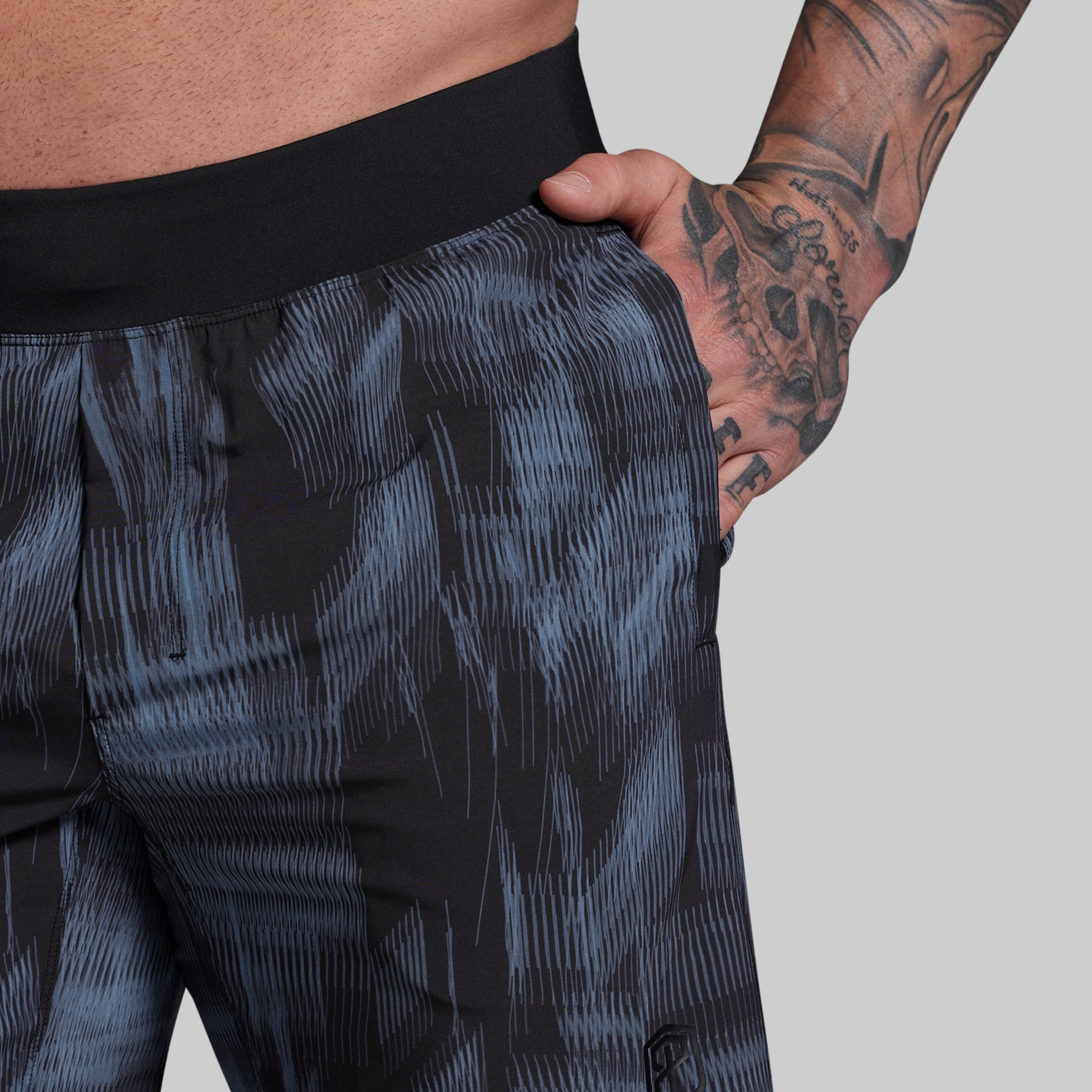 Person wearing dark patterned shorts with a tattooed arm on a plain background