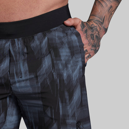 Person wearing dark patterned shorts with a tattooed arm on a plain background