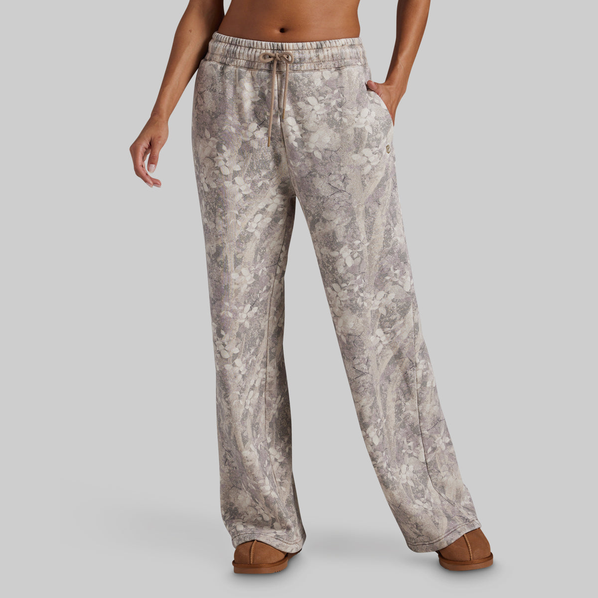 A model wearing wide-leg sweatpants with a beige and gray floral pattern, paired with brown slip-on shoes, shown from the waist down against a solid gray background.