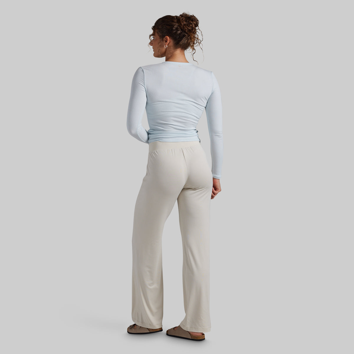 A woman shown from the back wearing a light blue long-sleeve top and cream wide-leg pants, standing against a plain light gray studio background.