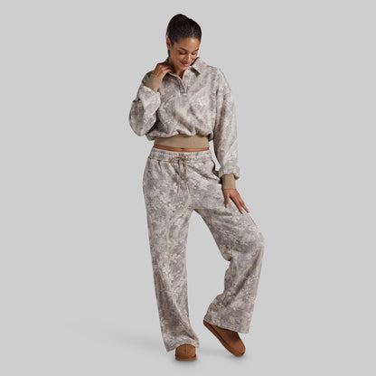 A woman modeling a matching two-piece loungewear set featuring a long-sleeve cropped polo sweatshirt and wide-leg sweatpants in a light gray and beige abstract pattern, styled with brown suede shoes against a solid gray background.