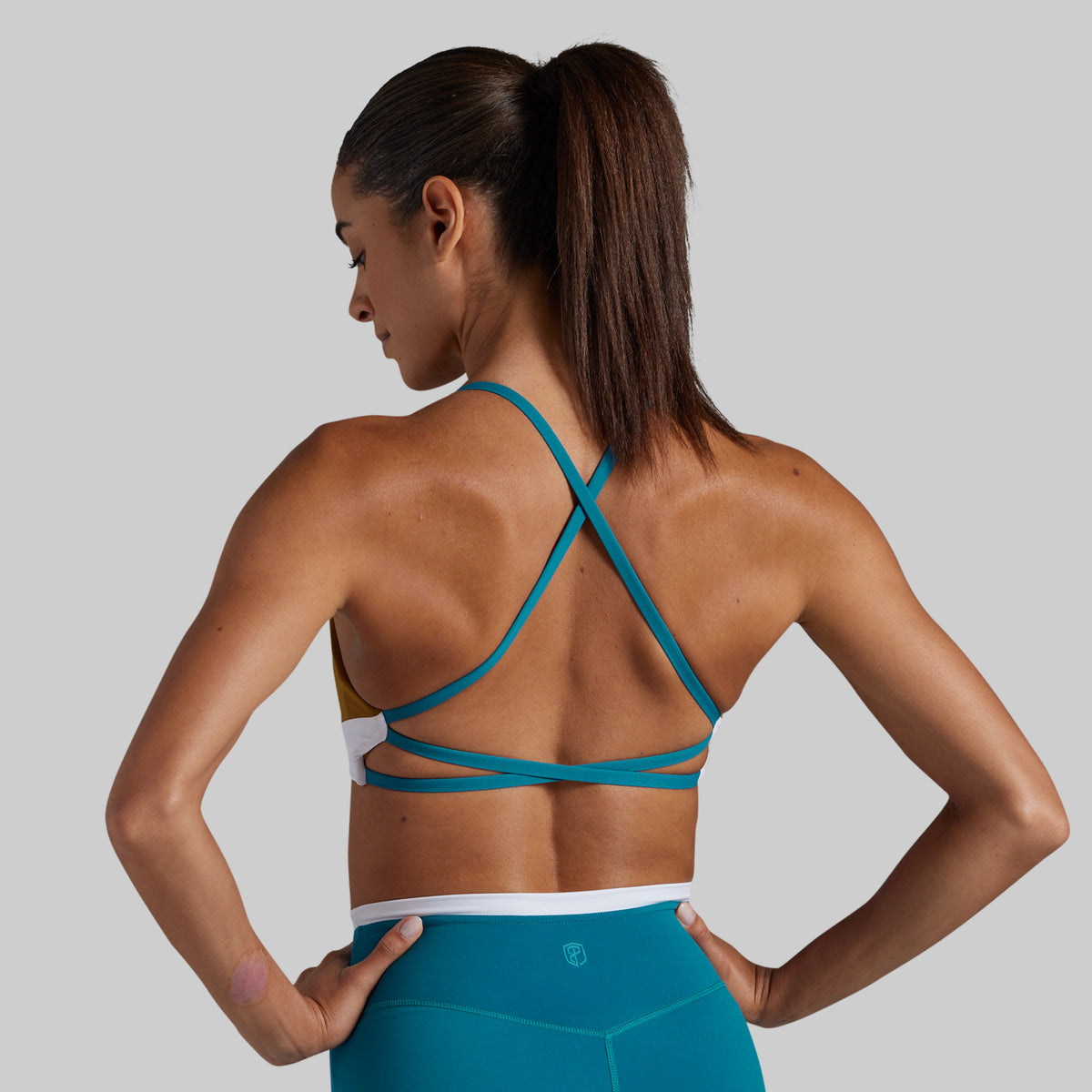 A woman shown from the back wearing a teal sports bra with crisscross straps and matching teal leggings, posed against a light gray studio background.