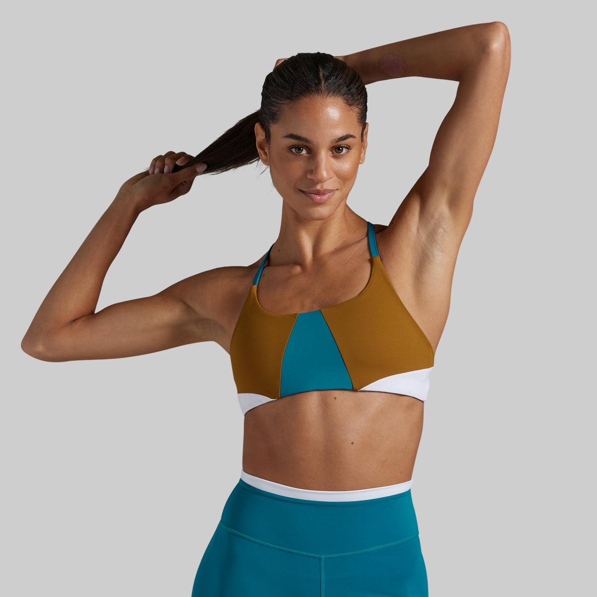 A woman modeling a color-blocked sports bra with a mustard yellow top, teal center panel, and white underband, paired with matching teal leggings, against a solid gray background.