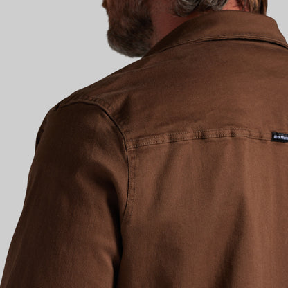 Close-up back view of a man wearing a brown overshirt or jacket, showing the collar, shoulder, and a small black brand tab on the upper right shoulder against a light gray background.