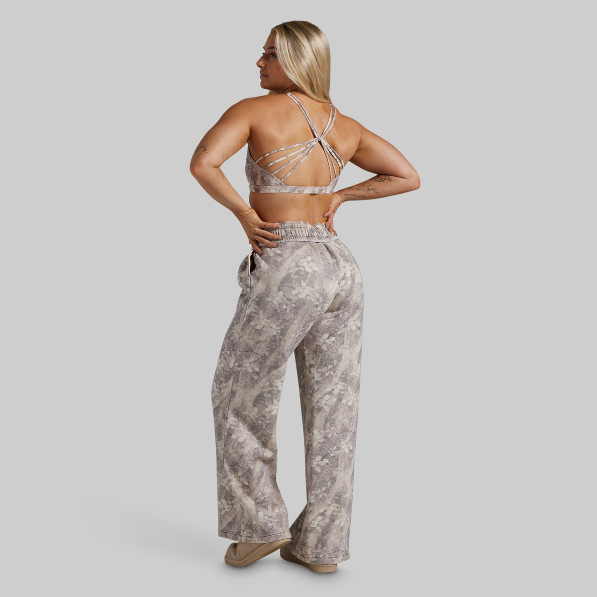 A woman is shown from the back wearing a matching two-piece lounge set consisting of a strappy sports bra and high-waisted, wide-leg pants in a gray and beige marble pattern, set against a solid gray background.