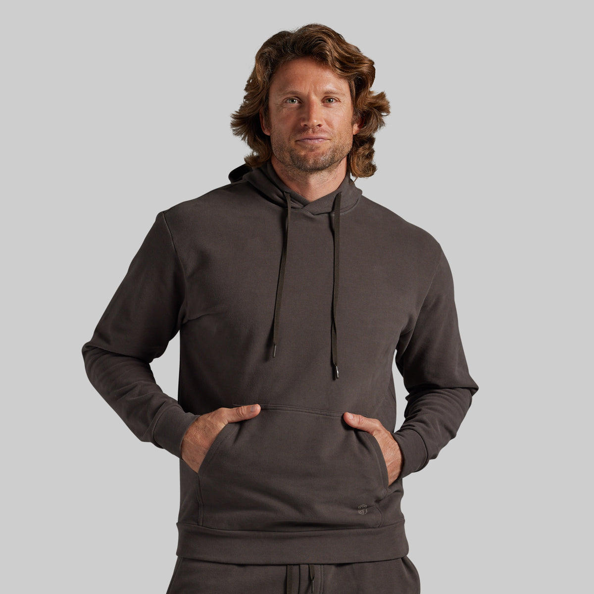 A man wearing a dark grey pullover hoodie with a drawstring hood and kangaroo pocket, standing against a solid gray background.