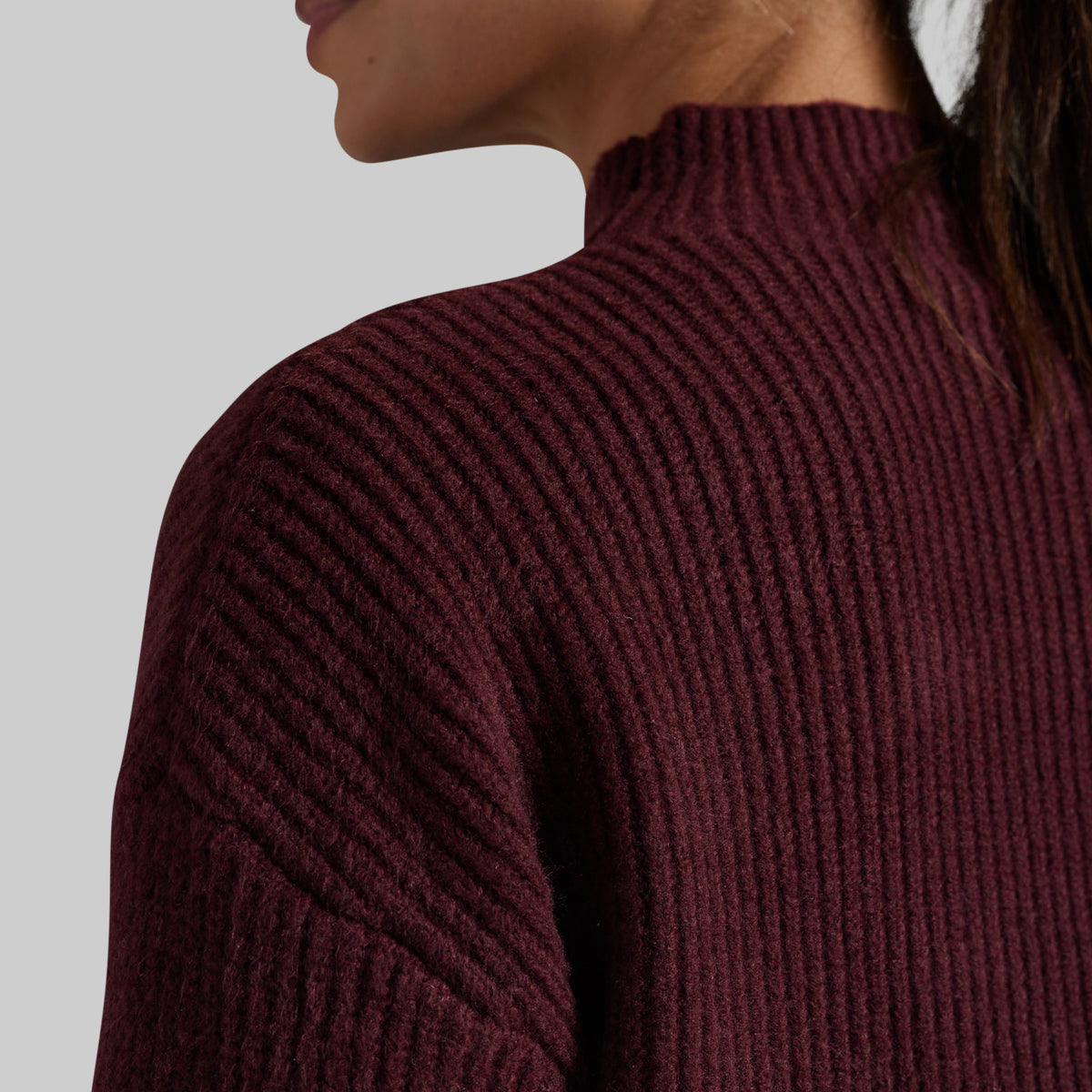 Close-up of a person wearing a ribbed knit mock neck sweater in deep burgundy against a light gray background.
