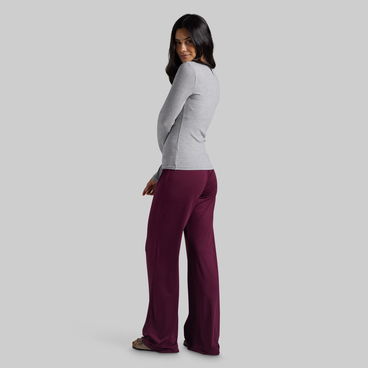 A woman modeling a light gray long-sleeve top and deep burgundy wide-leg pants, shown from a side-back angle against a solid light gray studio background.