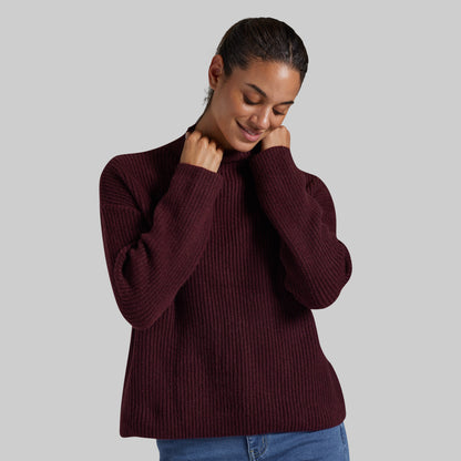 A woman wearing a ribbed knit mock neck sweater in deep burgundy, paired with blue jeans, against a solid gray studio background.