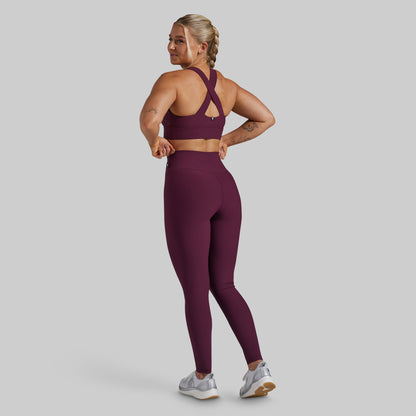 A woman modeling a matching burgundy activewear set, featuring a sports bra with a criss-cross back and high-waisted leggings, shown from a rear three-quarter angle against a solid gray background.