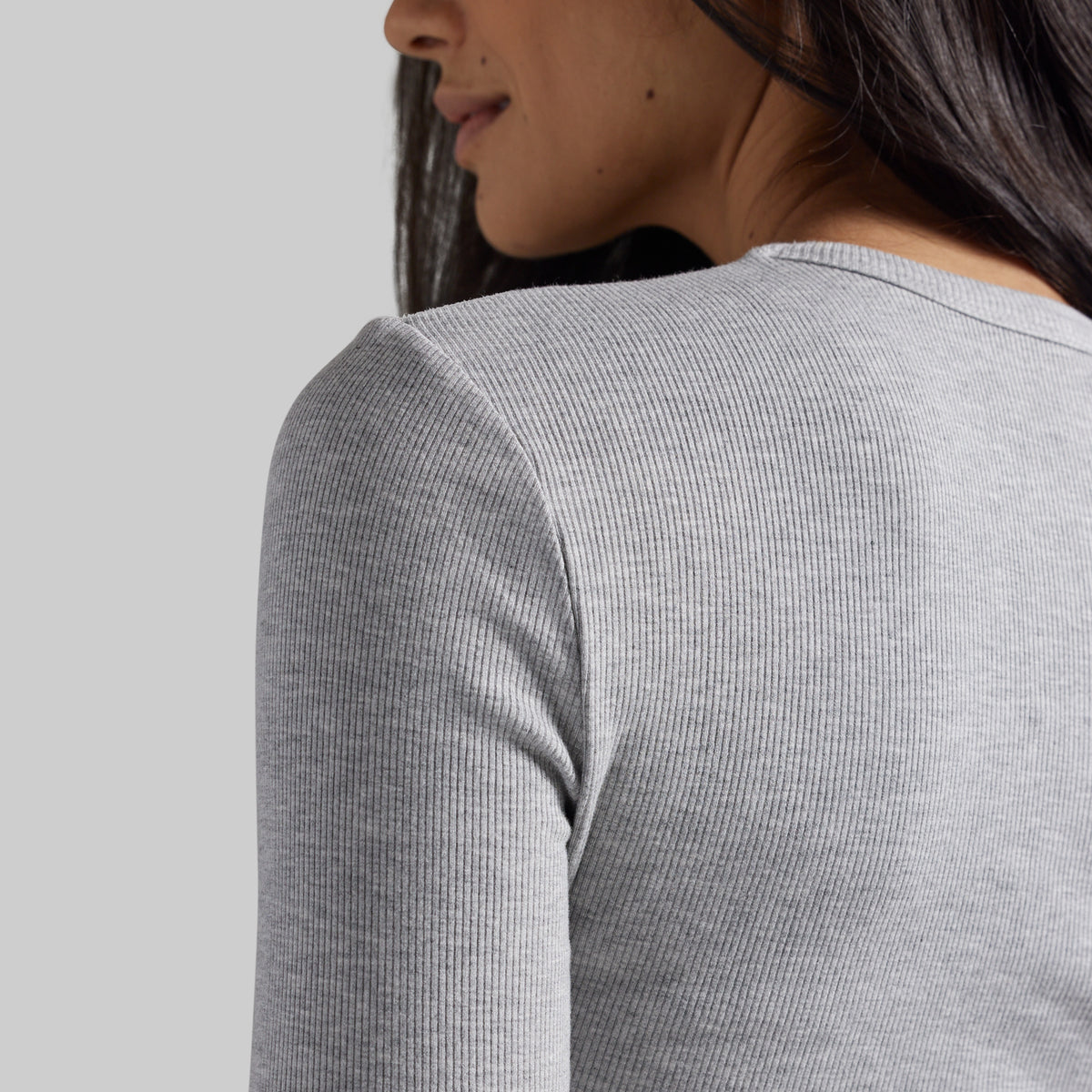 Close-up back view of a woman wearing a light heather gray ribbed long-sleeve top against a plain light gray background.