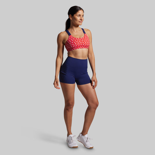 Woman wearing a red sports bra with white patterns and blue shorts on a gray background