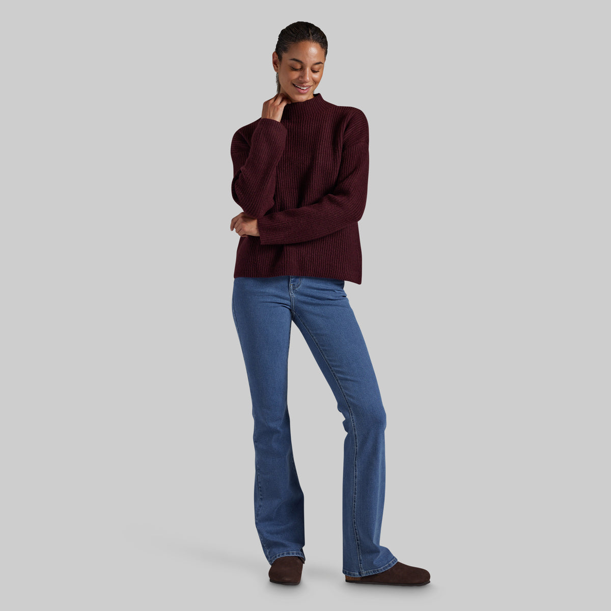 A woman modeling a ribbed long-sleeve turtleneck sweater in deep burgundy, paired with blue jeans and brown shoes, against a solid light gray background.