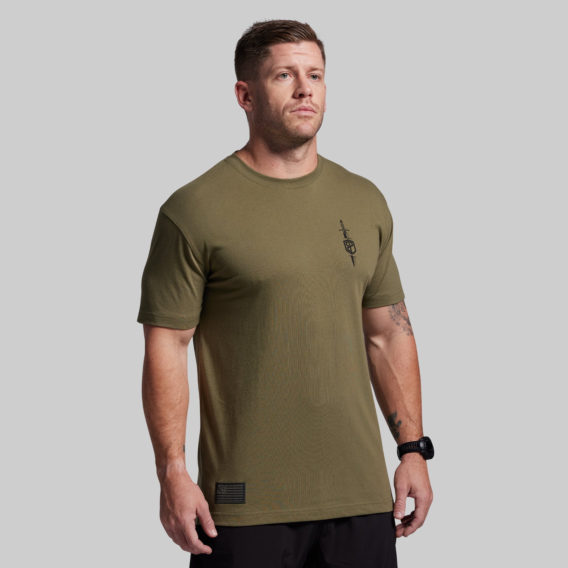 Man wearing an olive green t-shirt with a small logo on a gray background