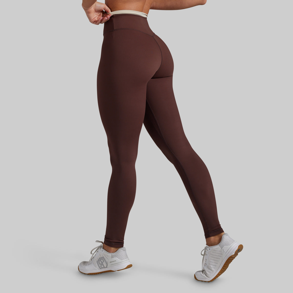 A model wearing high-waisted, form-fitting leggings in a dark brown color, shown from a rear three-quarter angle against a solid light gray background.