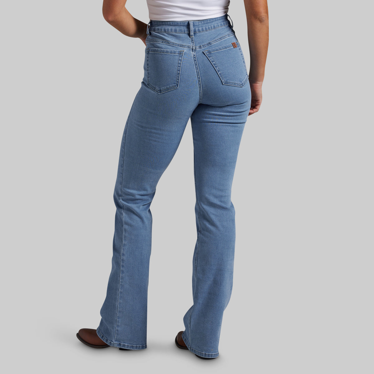 A person shown from the back wearing light blue denim jeans with a straight-to-bootcut leg, paired with a white top and brown shoes, against a solid gray studio background.