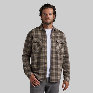 A man wearing a brown and beige plaid flannel shirt over a white t-shirt and gray pants, standing against a solid gray background.